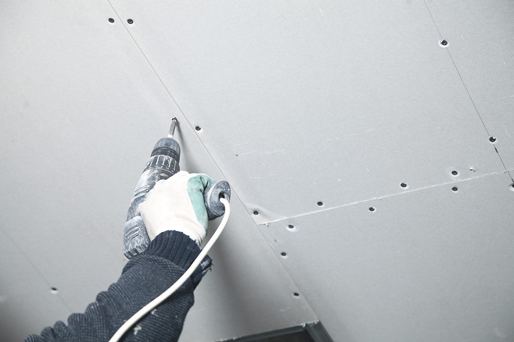 Worker with a drill screwdriver twists the screw into the drywall.