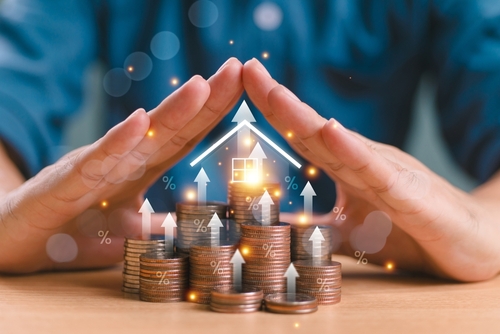 two hands covering a house with stacked coins in front.